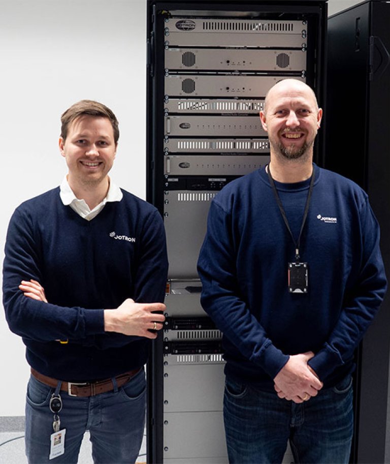 Two Jotron employees standing in front of a radio rack.