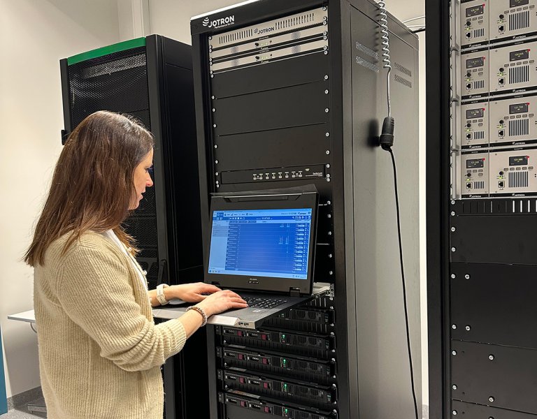 A woman standing in front of a radio equipment rack while monitoring information displayed on a PC screen.